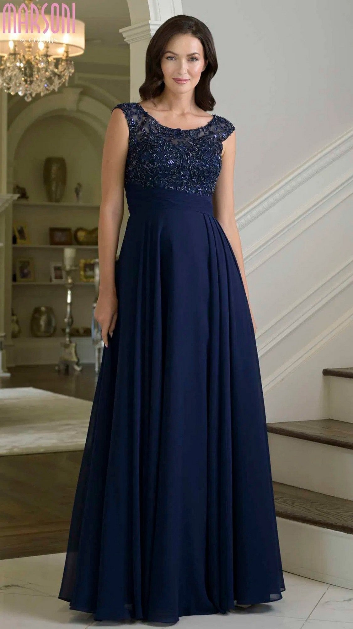 A woman stands indoors wearing the Marsoni M335 long formal dress in navy, featuring beaded lace, cap sleeves, and a floor-length chiffon A-line gown—perfect for any special occasion.